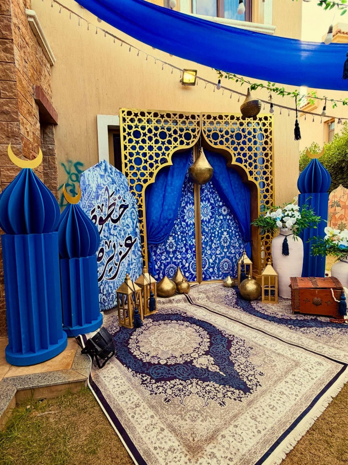 Festive blue and gold Eid decorations with lanterns, rug, and geometric screen outdoors.