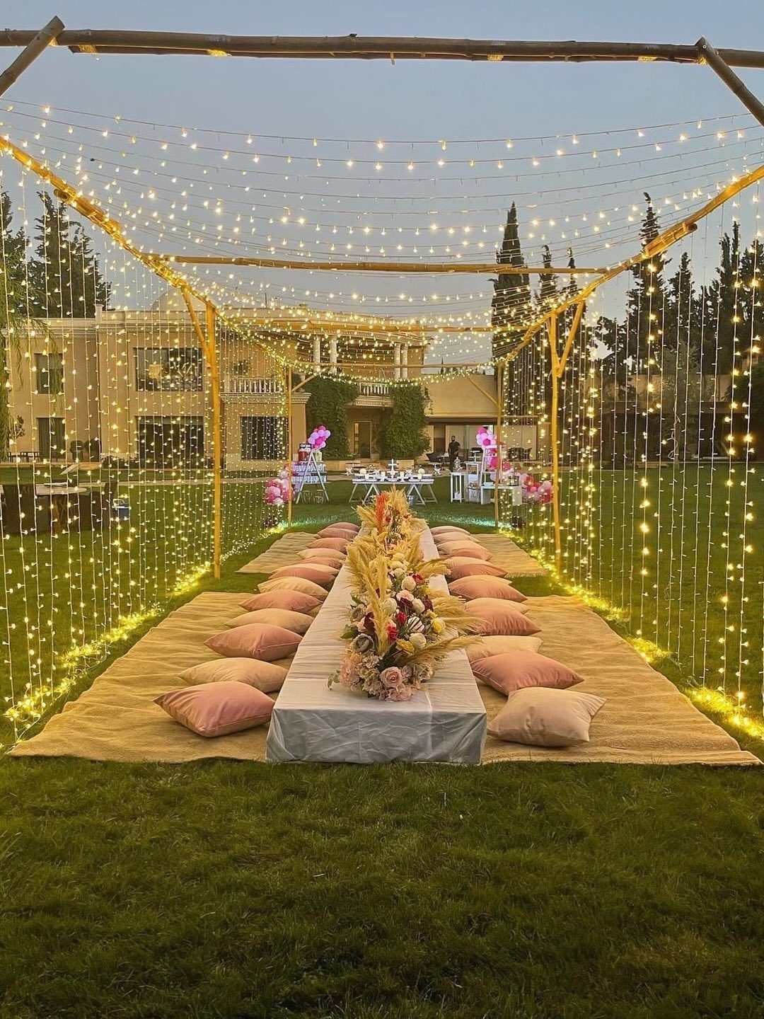 Outdoor dining setup with fairy lights, pink cushions, and floral centerpieces on grass.