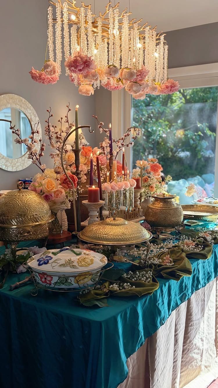 Teal tablecloth with gold platters, fresh flowers, and candles under a floral chandelier.