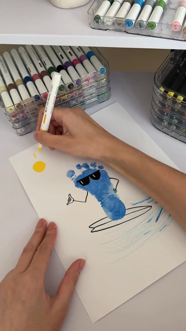 Hands drawing a yellow sun next to a blue footprint surfing character.