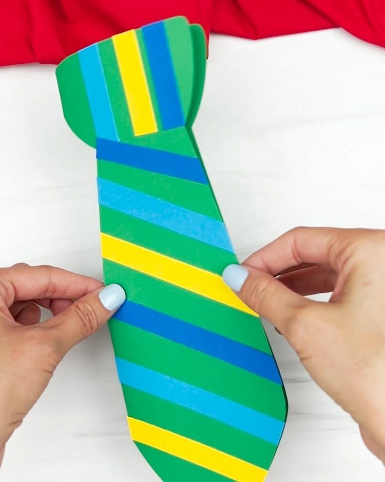 Hands holding a colorful striped paper necktie craft on a white surface