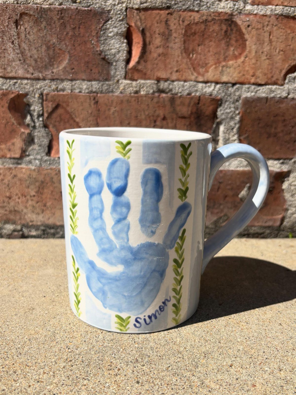 White ceramic mug with a blue child's handprint and the name Simon.