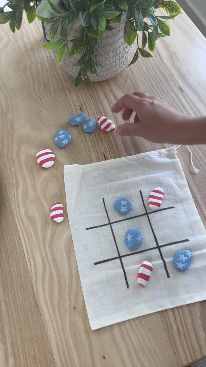 Hand playing tic-tac-toe with red, white, and blue painted stones on fabric