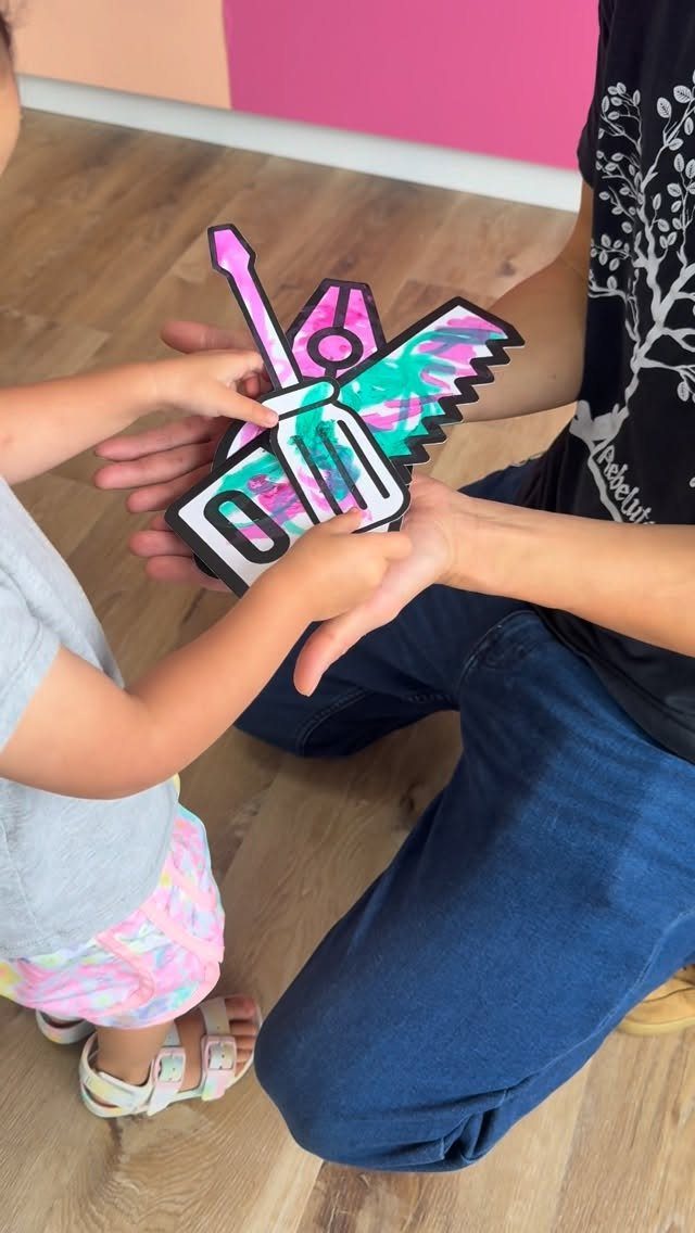 Child handing a colorful paper tool craft to a man on a wooden floor