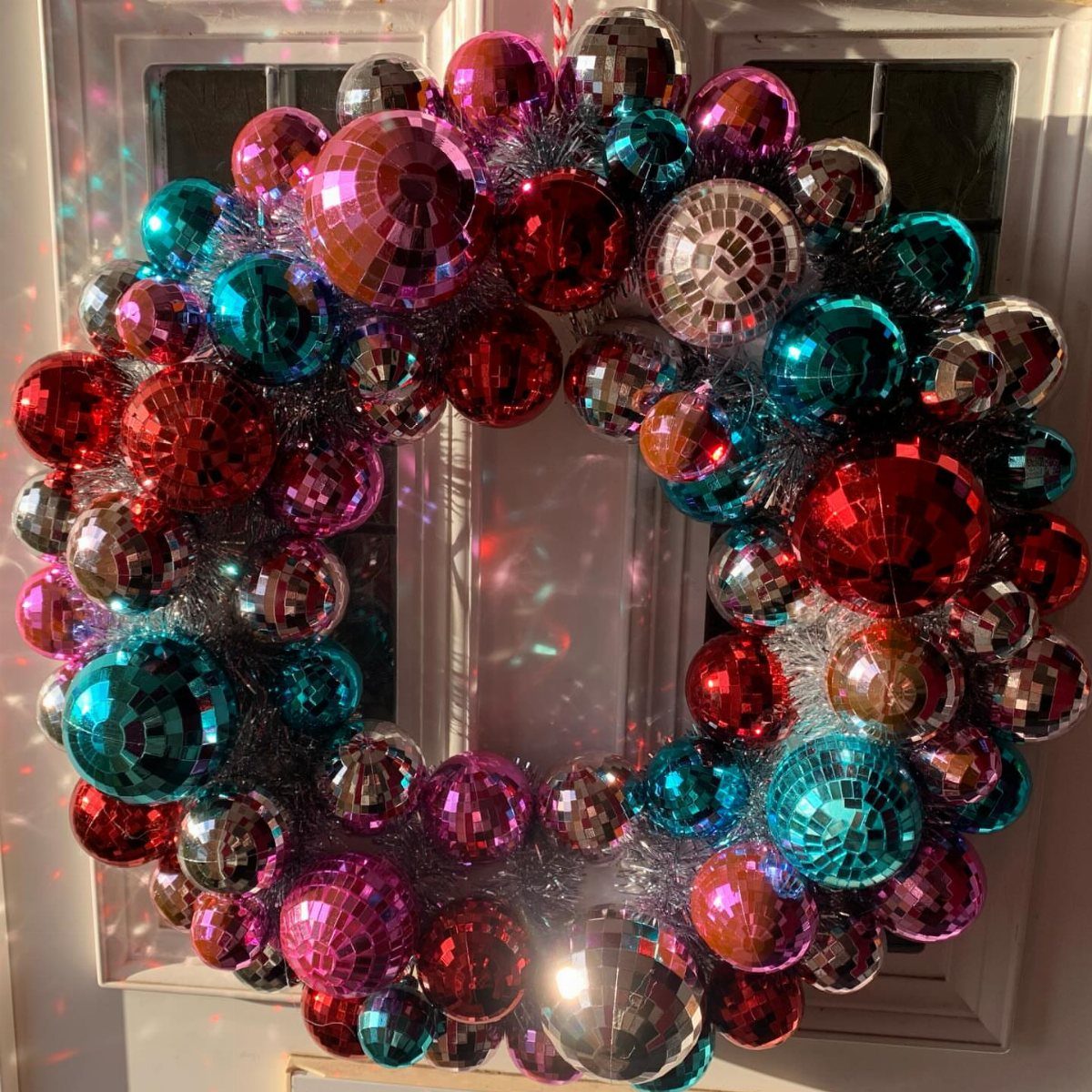 Colorful disco ball wreath hanging on a white front door with light reflections.