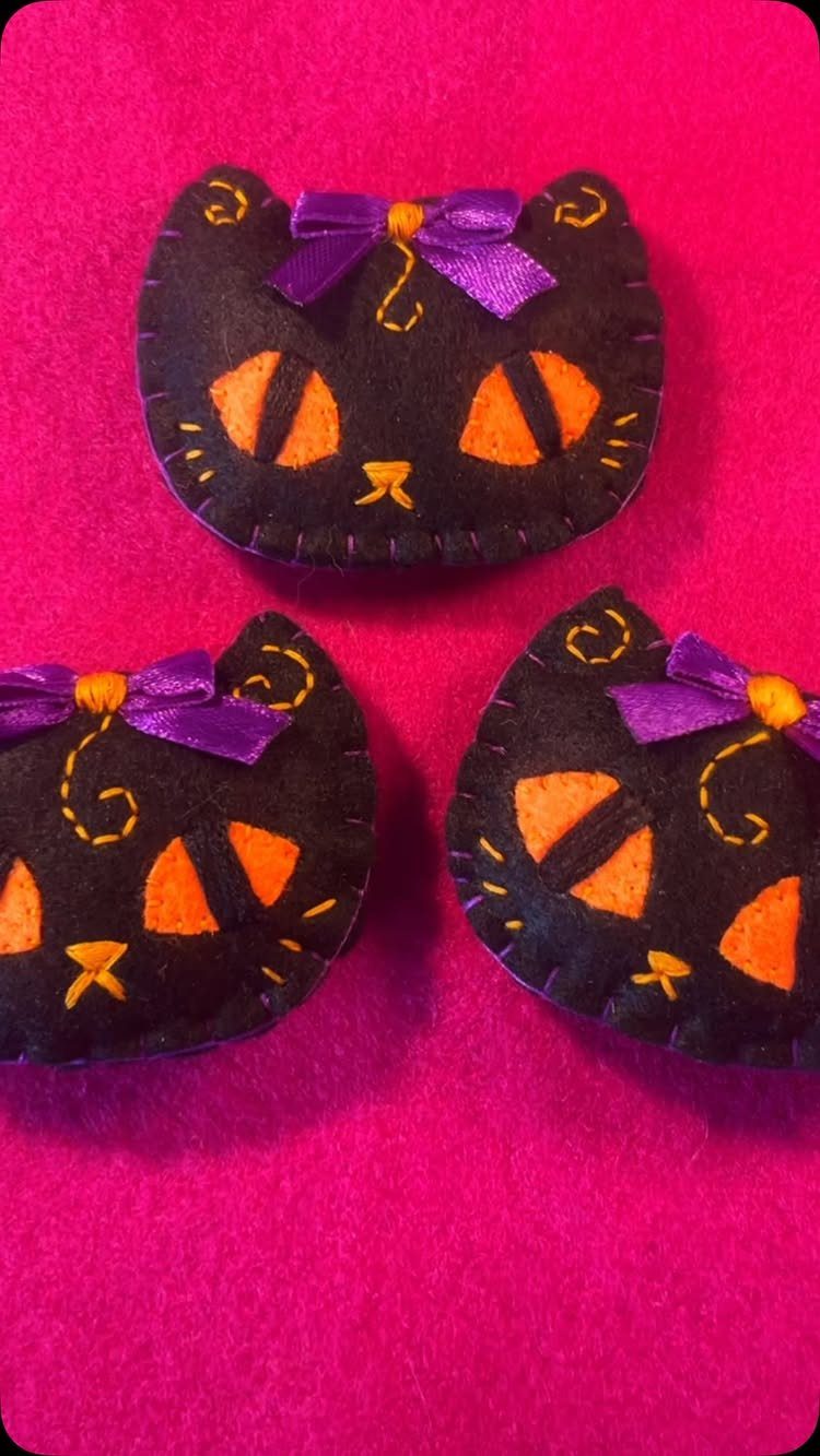 Three black felt cat faces with orange eyes and purple bows on pink felt.