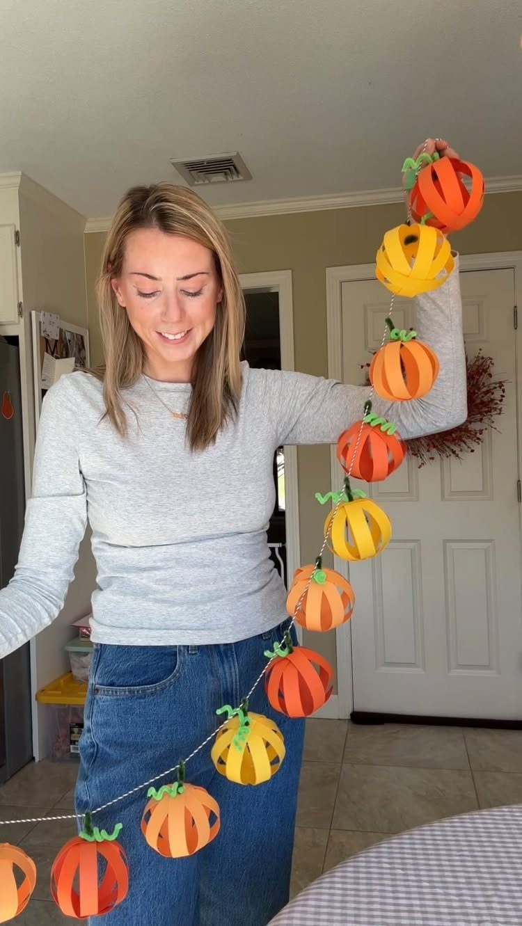 Woman holding a handmade garland of orange and yellow paper pumpkins indoors.