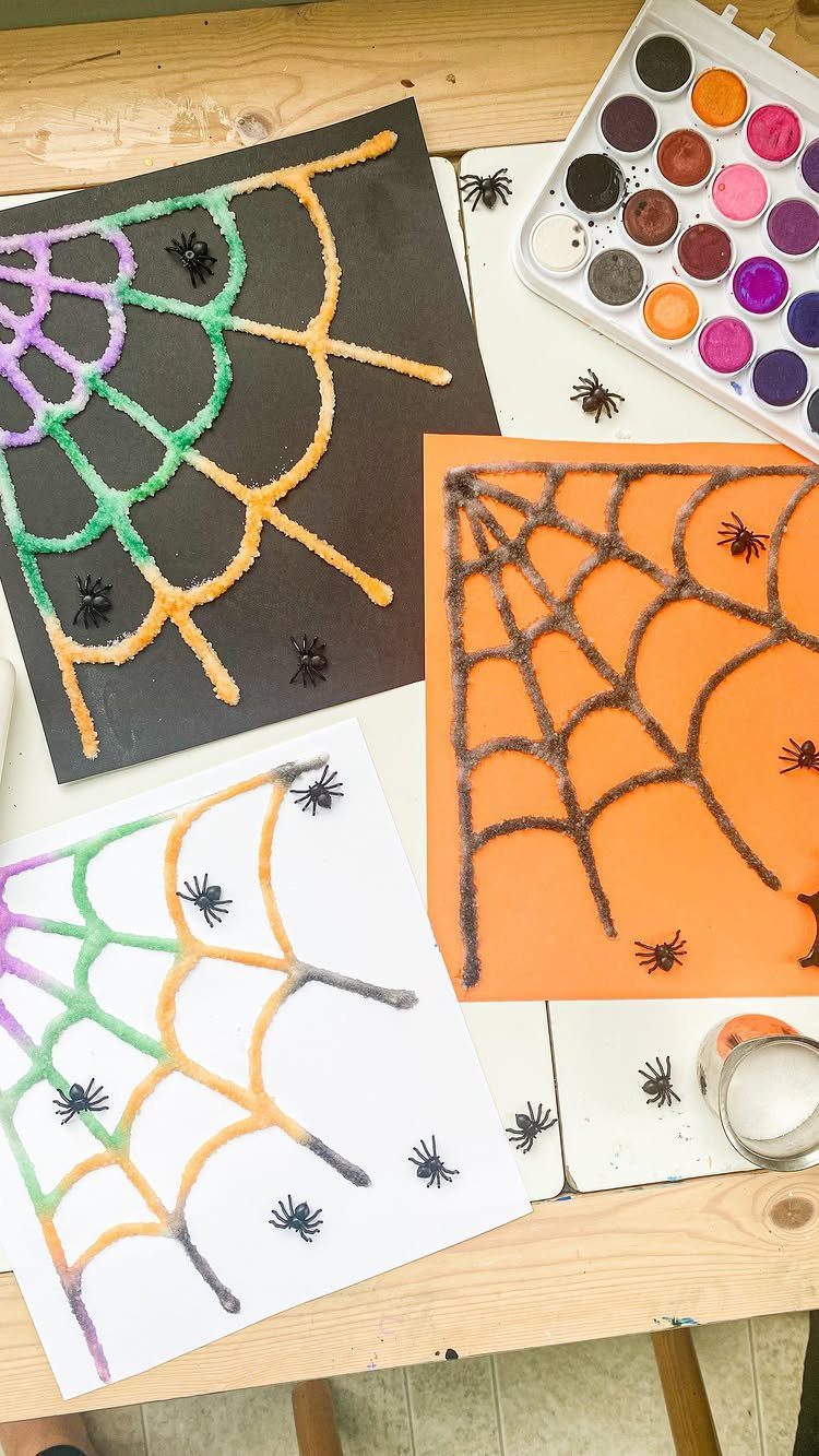 Colorful salt painted spiderwebs on black, orange, and white paper with plastic spiders.