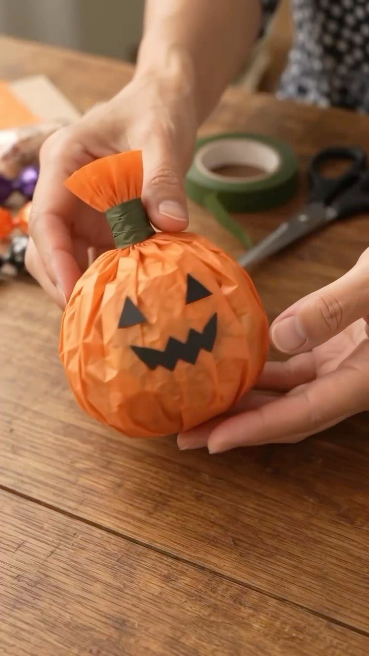 Orange tissue paper pumpkin with a black face held by two hands.