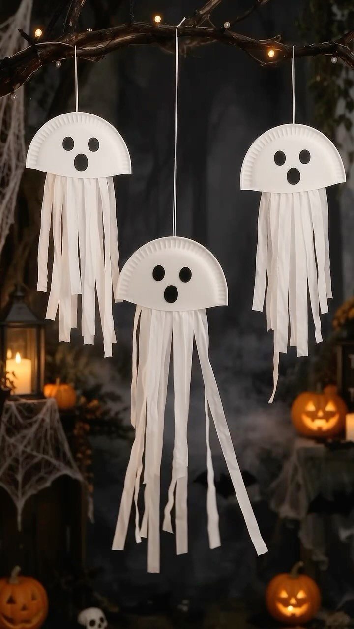 Three white paper plate ghosts hanging from a branch among glowing pumpkins.