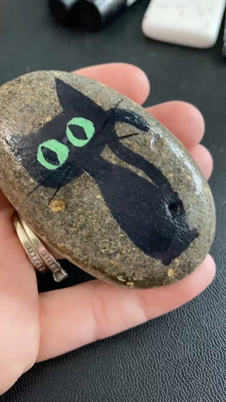 Painted river stone featuring a black cat with large green eyes held in hand