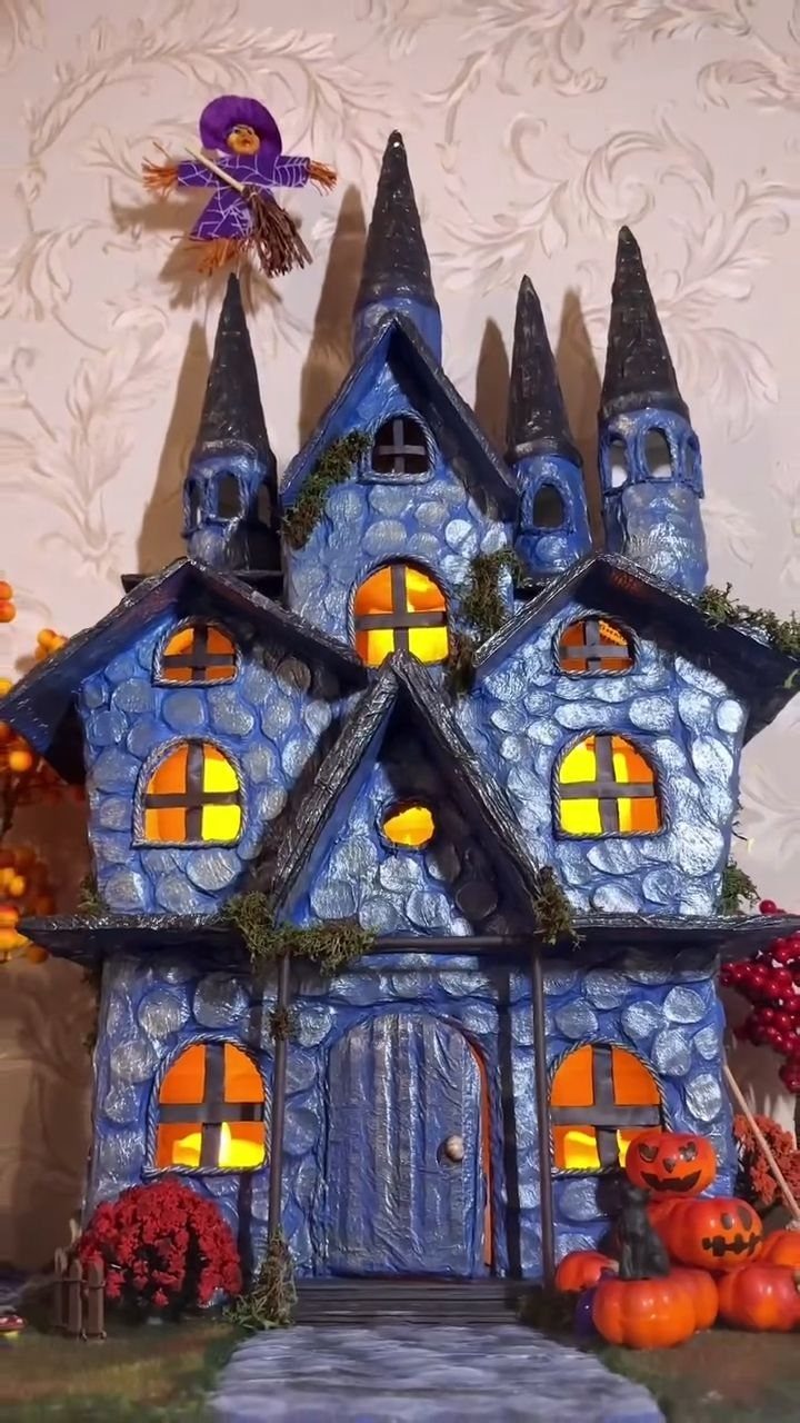 Handmade blue miniature haunted house with glowing windows and small orange pumpkins.