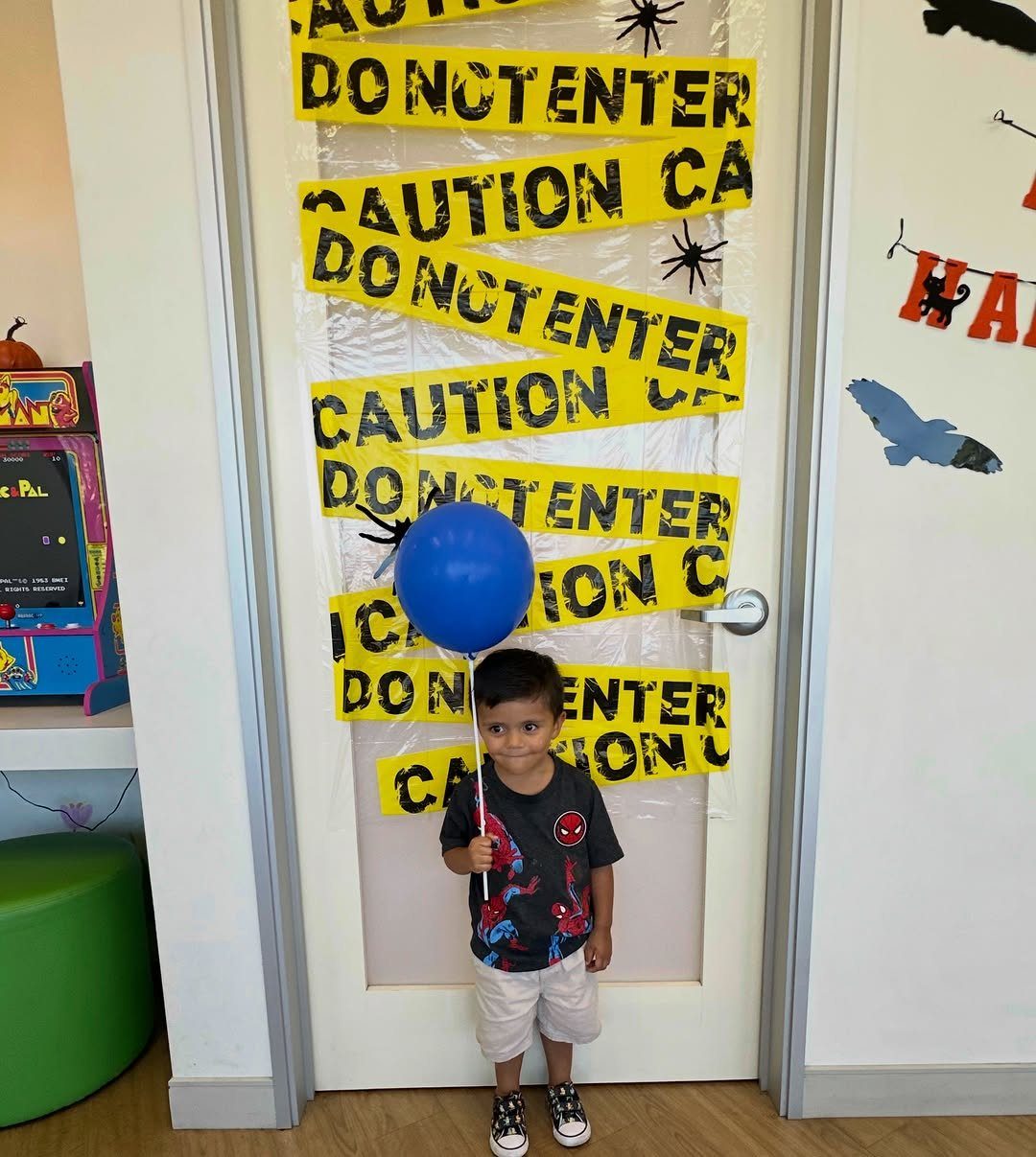 Boy with blue balloon standing before a door decorated with caution tape and spiders