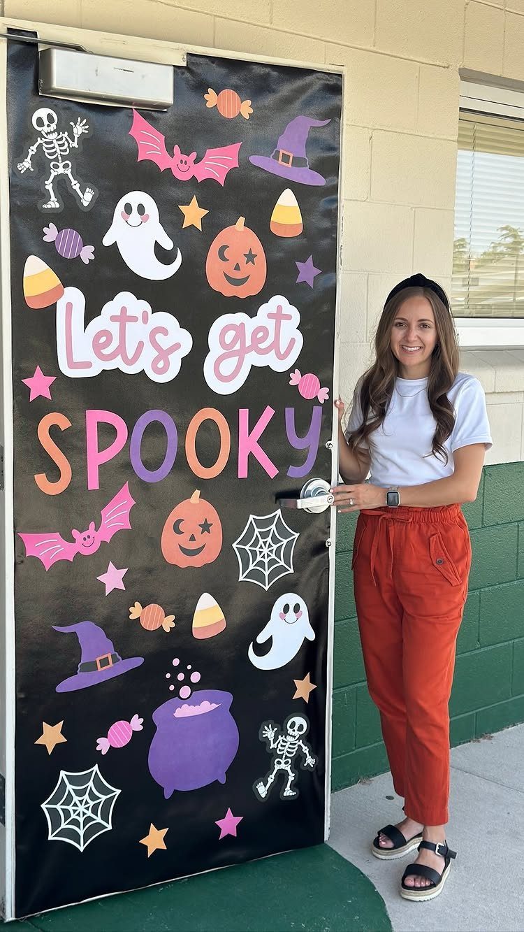 Woman standing beside a black classroom door decorated with colorful Halloween cutouts and text.