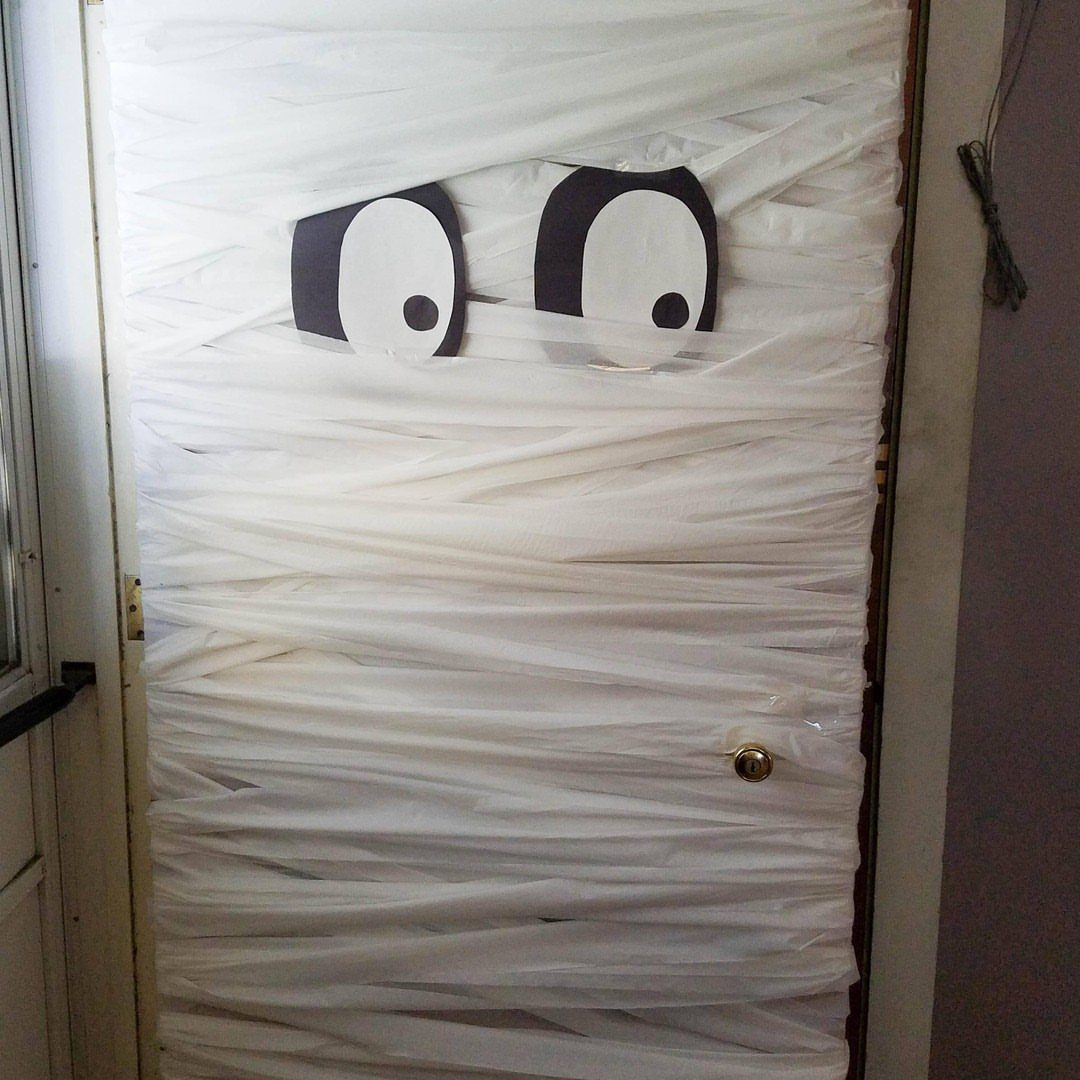 Mummy door decoration wrapped in white fabric strips with large cartoon eyes.