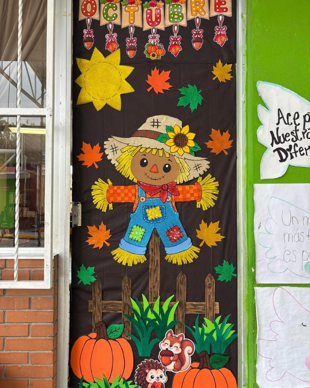 Paper scarecrow decoration on a classroom door with pumpkins and autumn leaves.