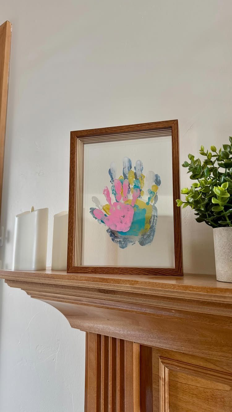 Framed colorful child's handprint art sitting on a wooden mantel with a plant.