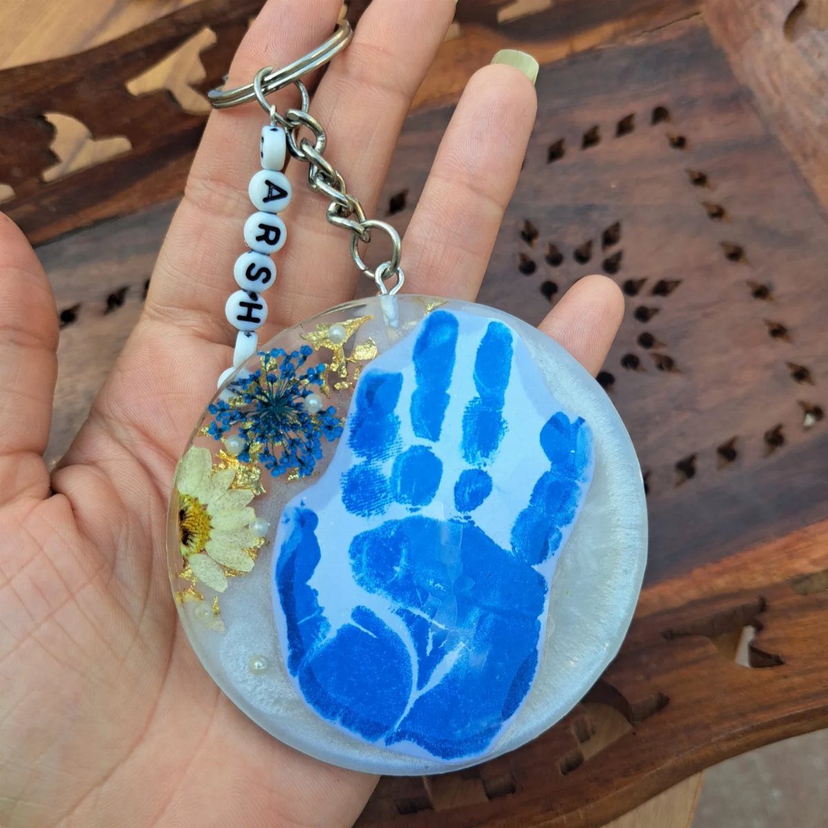 Circular resin keychain featuring a blue handprint, dried flowers, and beaded name strap.