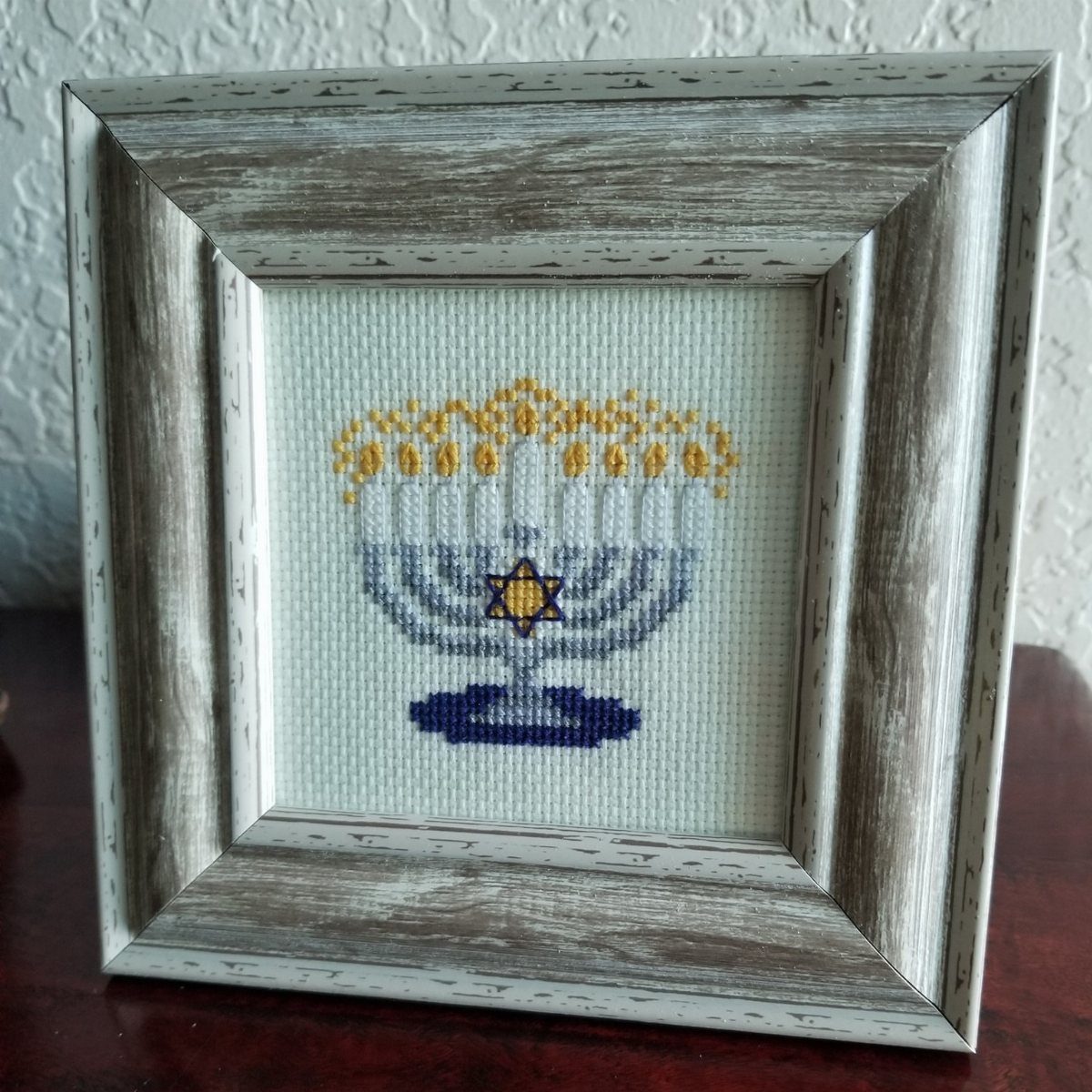 Cross-stitched menorah with blue and yellow threads in a rustic wooden frame.