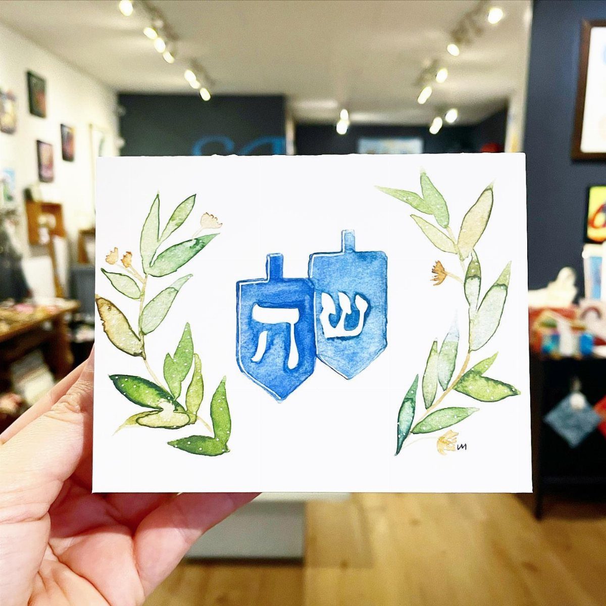 Hand holding a watercolor greeting card with blue dreidels and green leaves.