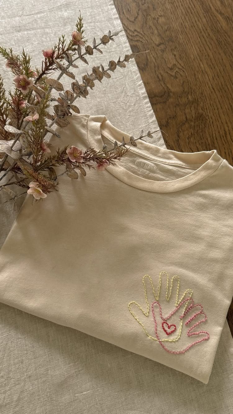 Cream t-shirt with embroidered yellow and pink handprints and a small red heart.