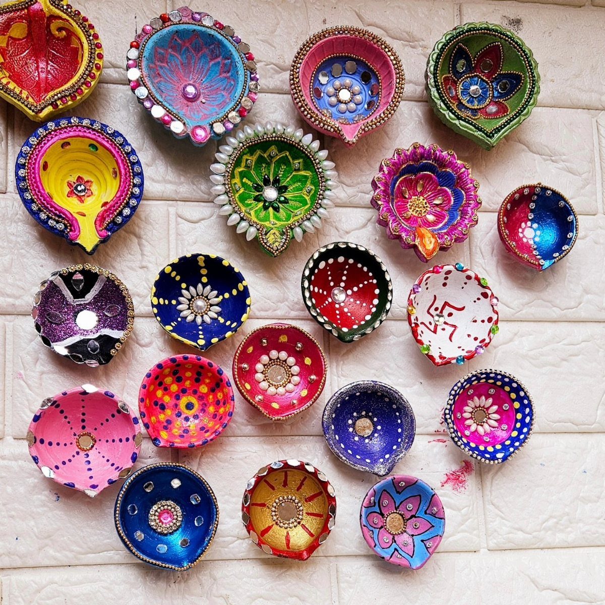 Colorful hand-painted clay diyas with mirrors and glitter on a white brick background
