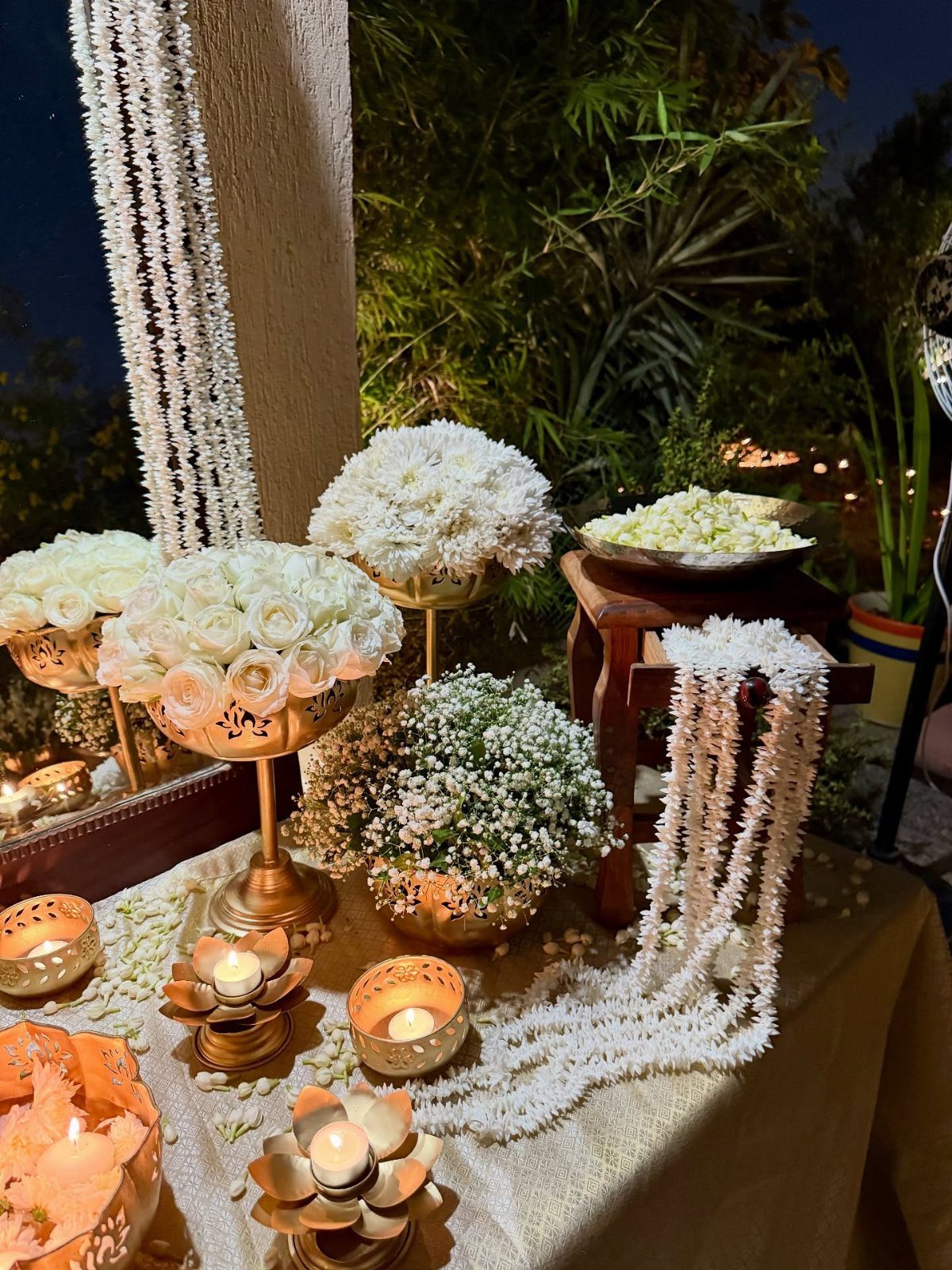 White flowers in gold vases and bowls with lit candles on a table.