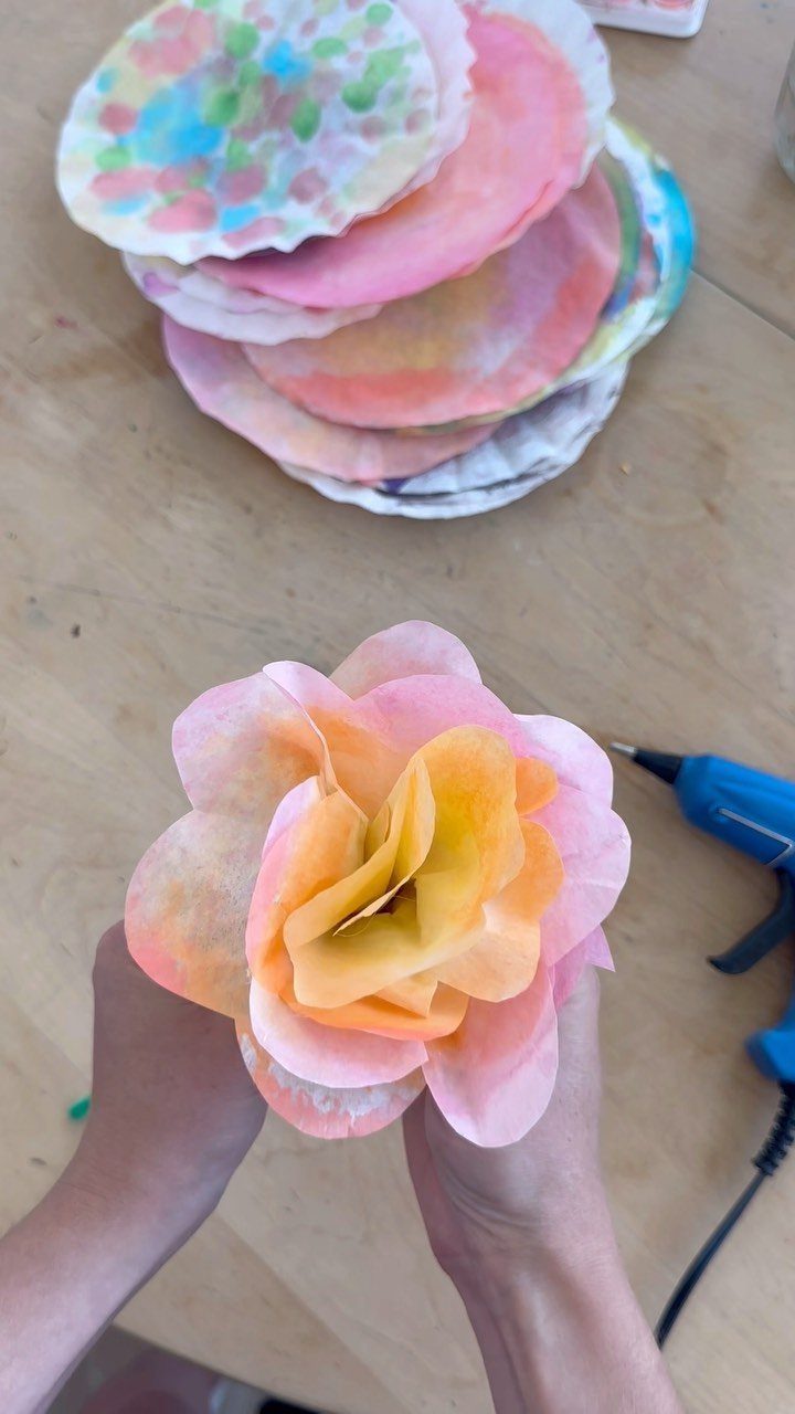 Hands holding a pink and orange coffee filter flower on a wooden table.