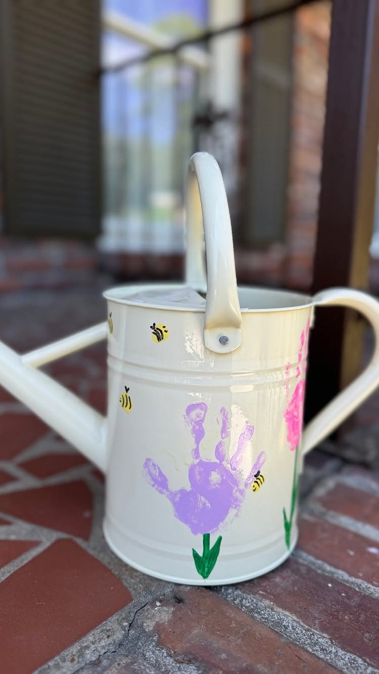 White watering can decorated with a purple handprint and painted flowers on bricks.