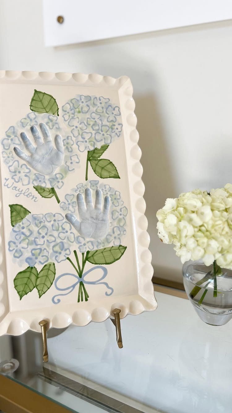 Ceramic tray with blue hydrangea flowers and infant handprints, displayed on a stand.