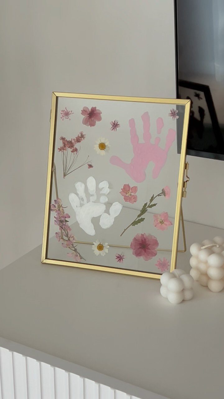 Baby handprints and dried flowers pressed inside a gold glass frame on a table.