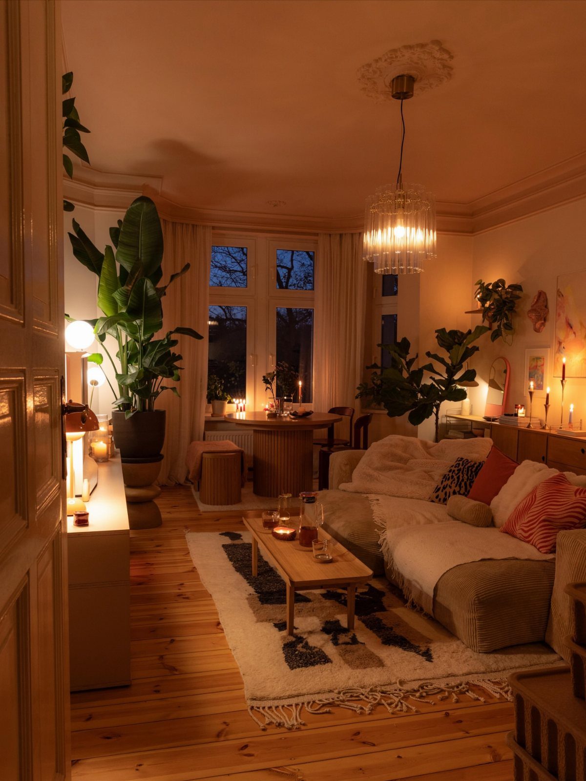 Cozy living room with warm lighting, lit candles, and large indoor green plants.