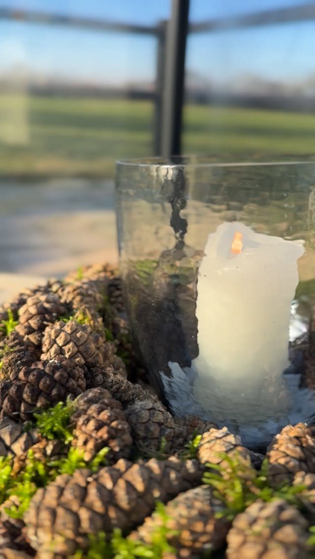Burning white pillar candle in glass surrounded by pinecones and green moss
