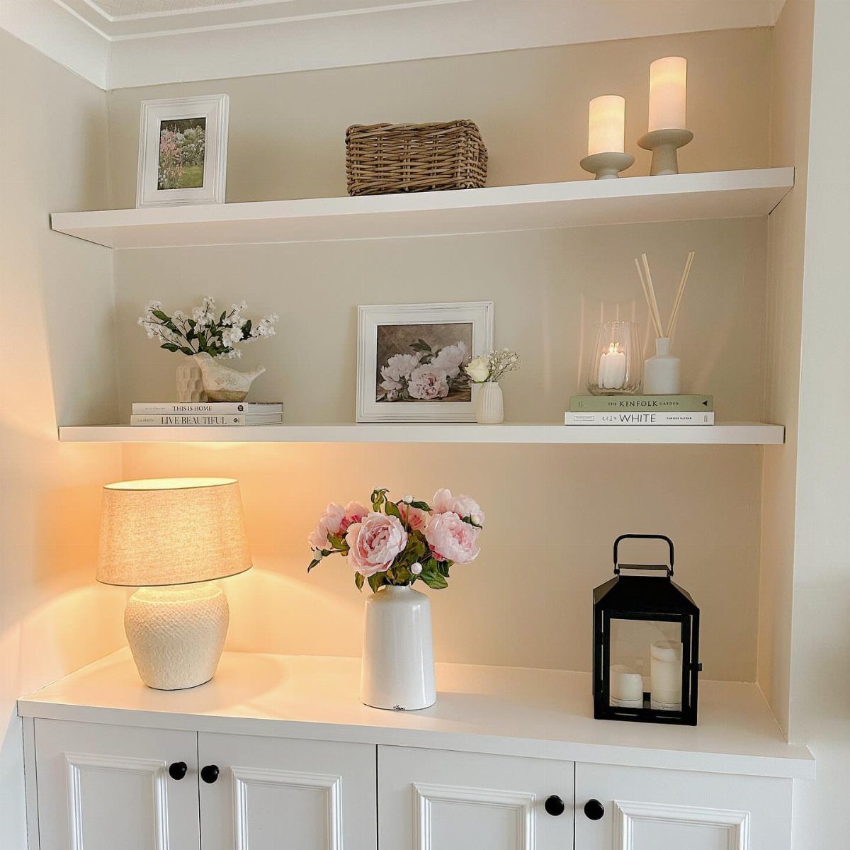 White built-in shelves decorated with pink flowers, candles, framed art, and a lamp.