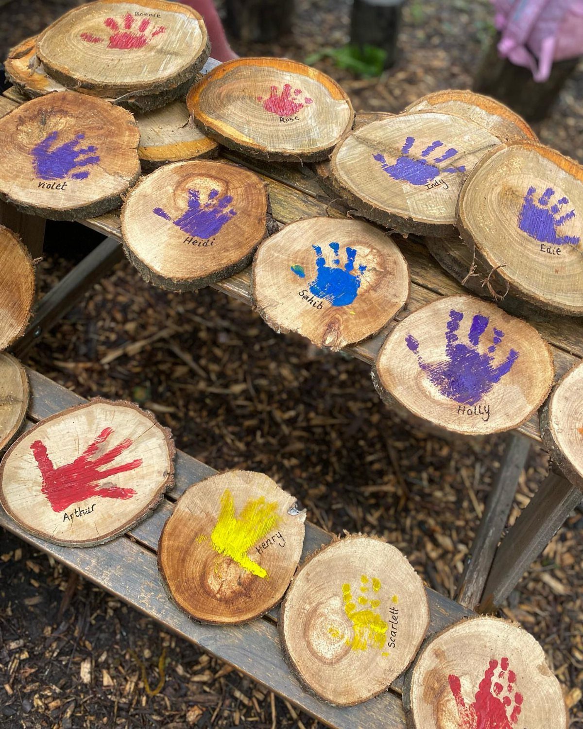 Wooden slices with colorful children's handprints and names resting on a wooden bench.