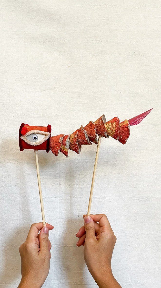Red and gold egg carton dragon puppet held up by two wooden skewers.