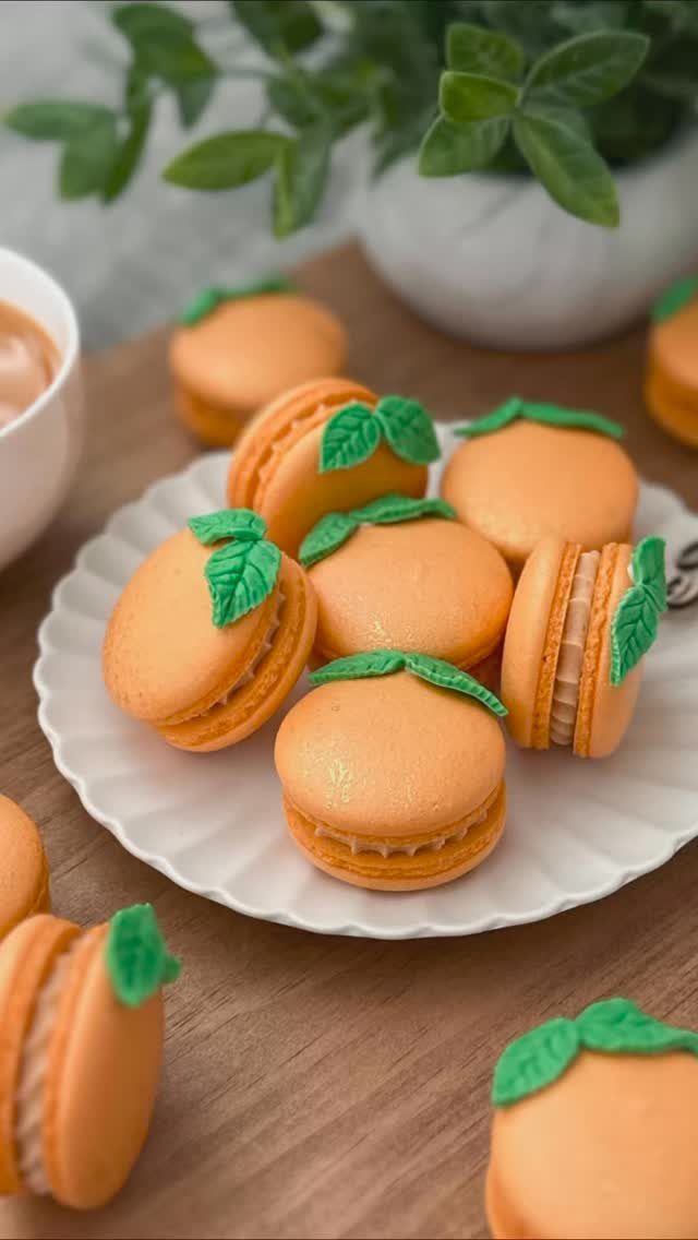 Orange macarons shaped like mandarin oranges with green leaves on a white plate.