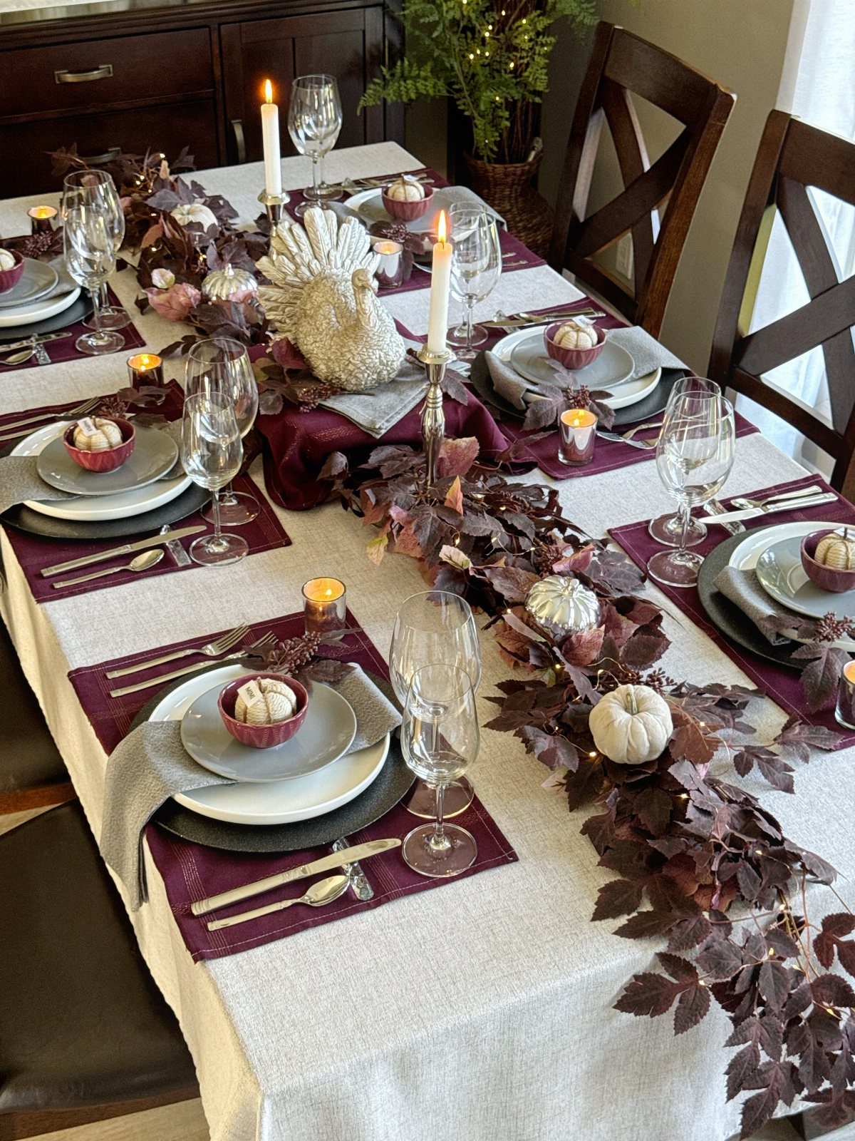 Thanksgiving table setting with burgundy accents, white pumpkins, and a leaf garland centerpiece.