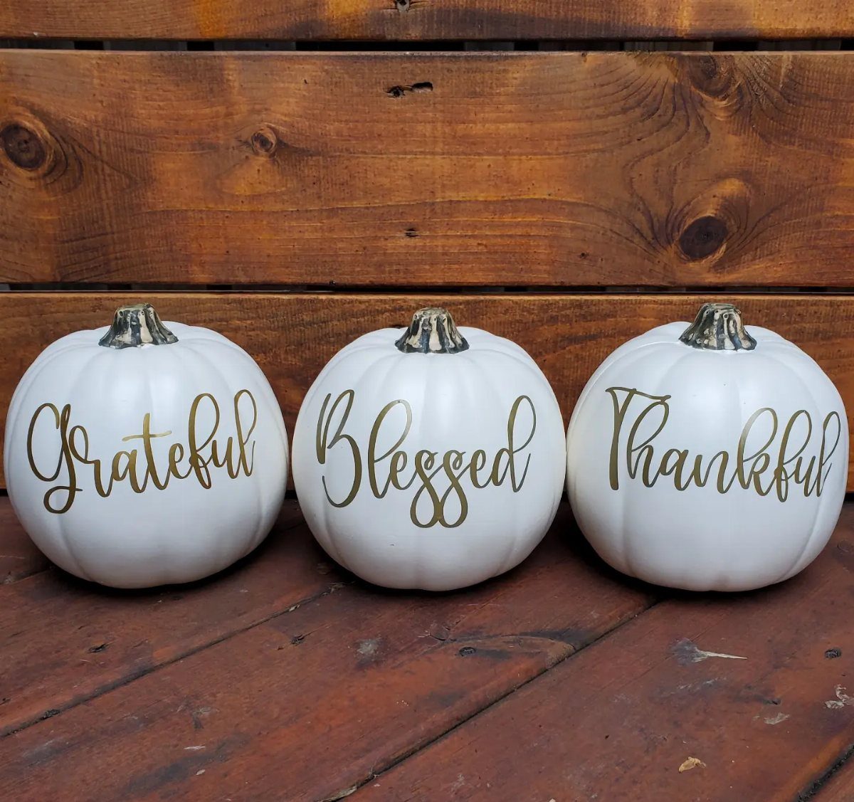 Three white pumpkins with gold lettering on a rustic wooden background.
