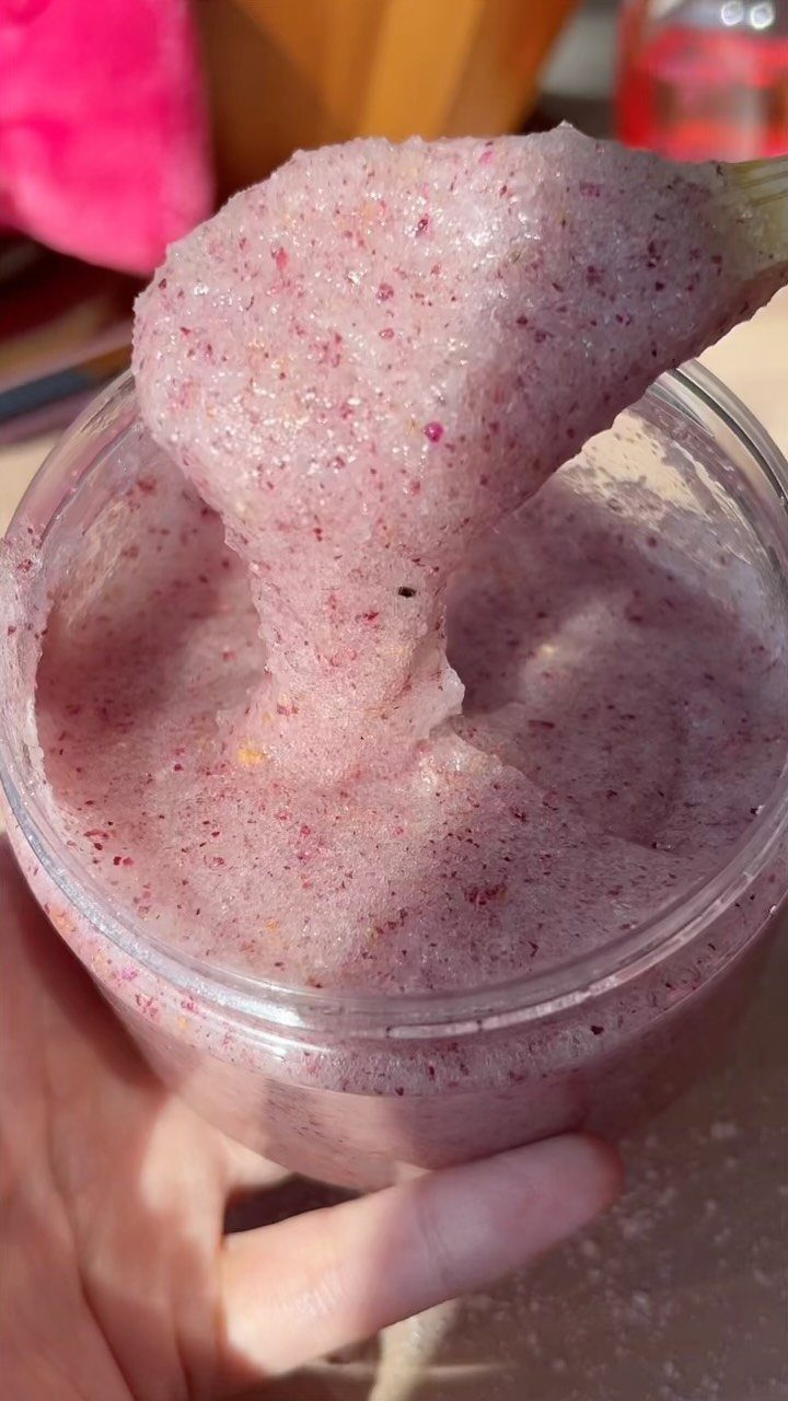 Pink sugar scrub with red flecks being scooped from a clear plastic jar