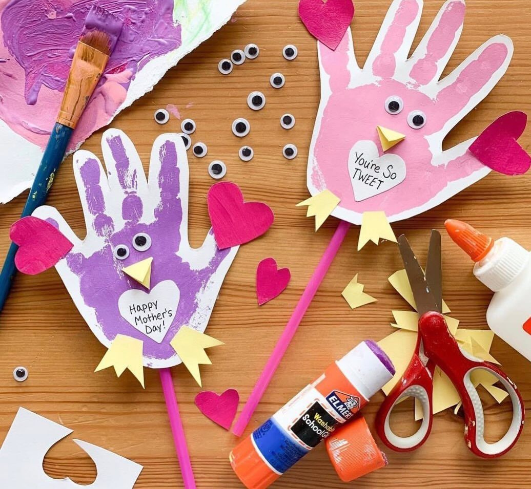 Handprint bird crafts with hearts and craft supplies on a wooden surface.