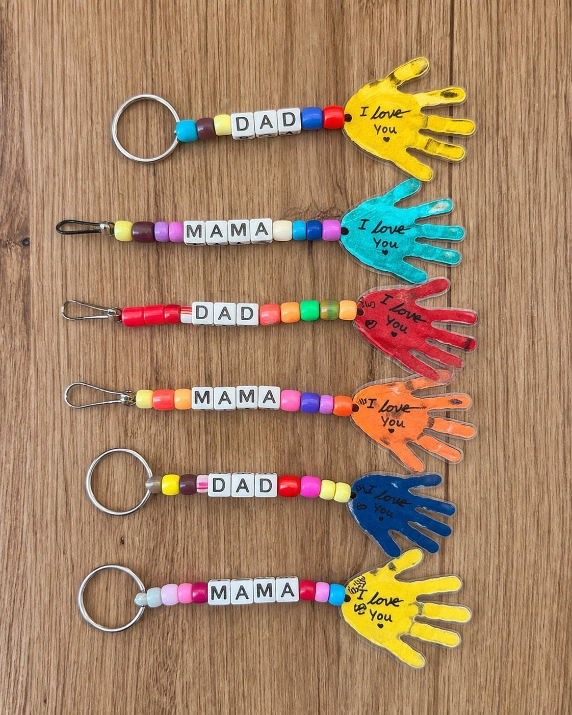Six colorful handprint keychains with beads spelling Mama and Dad on wood.