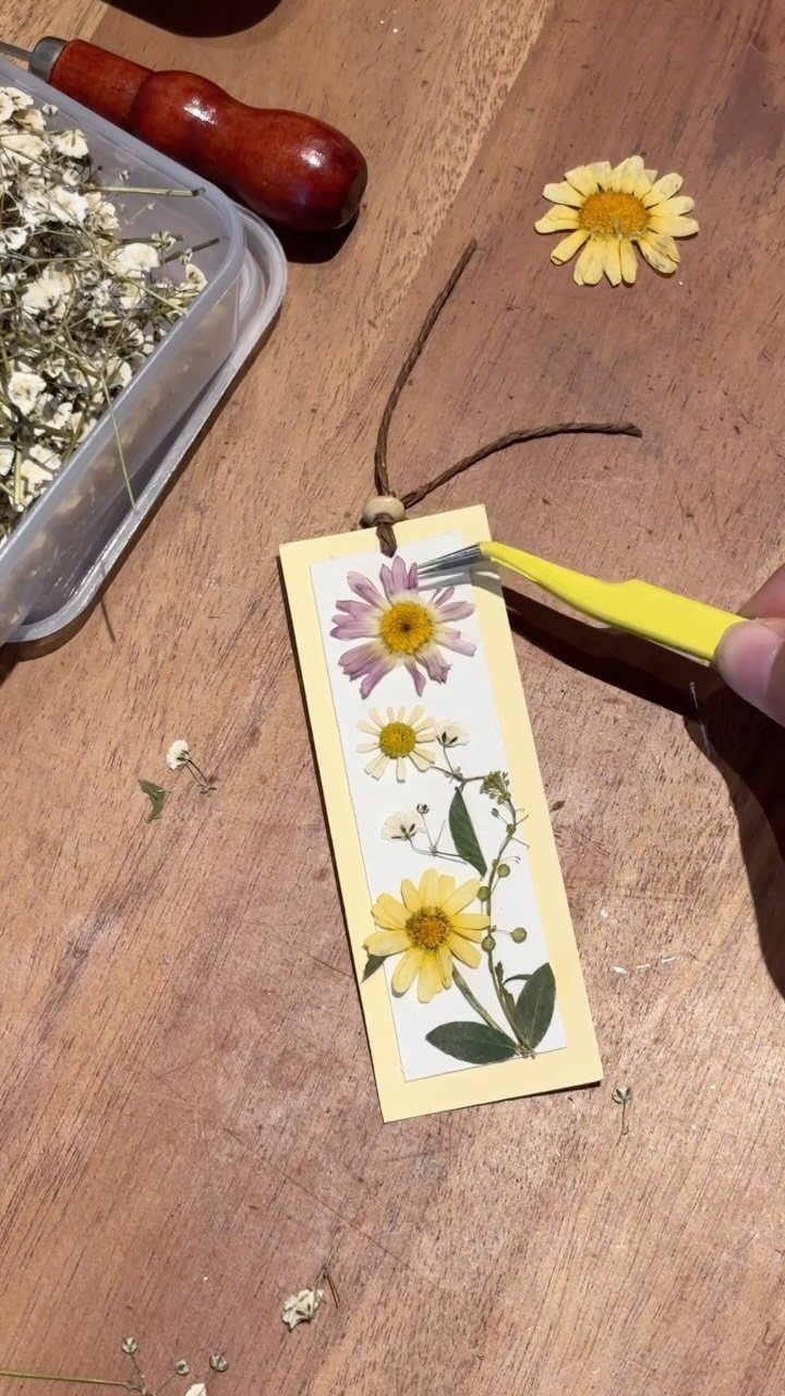 Pressed flower bookmark being assembled with tweezers on a wooden table.