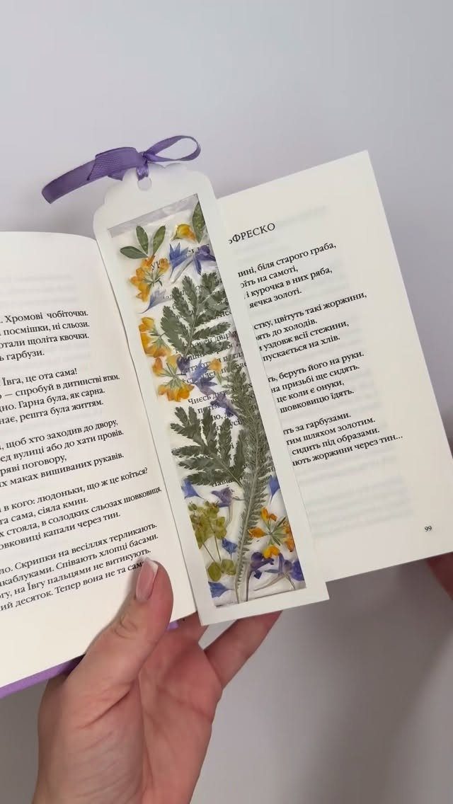 Pressed flower bookmark with a purple ribbon inside an open book held by hand