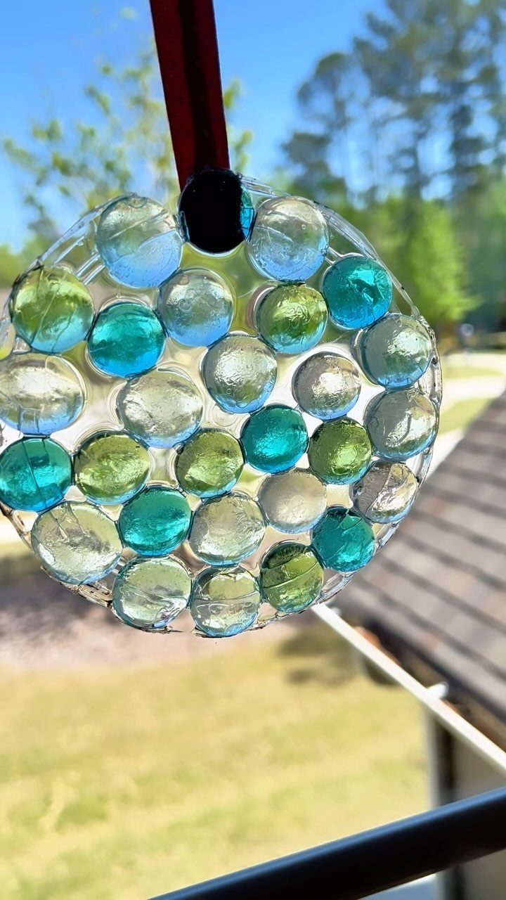 Circular resin suncatcher with colorful glass marbles hanging by a red ribbon outdoors.