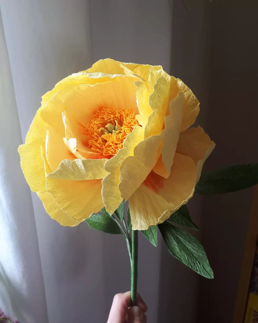 Yellow crepe paper flower with orange center and green stem held by hand.