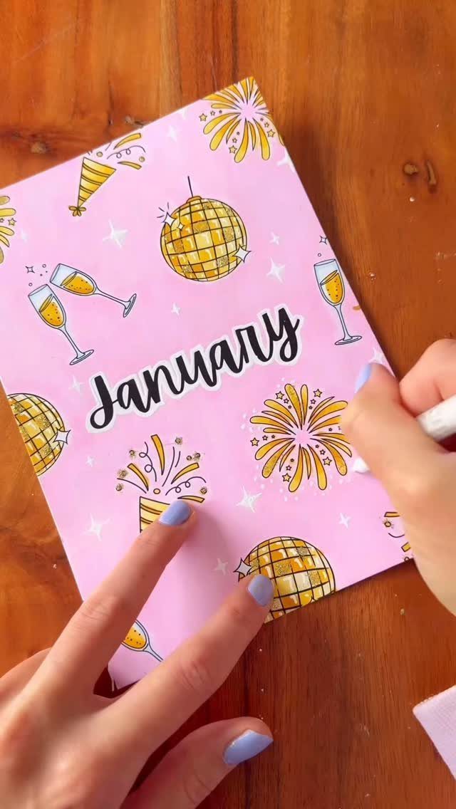 Hands coloring a pink January page with disco balls and fireworks on wood.