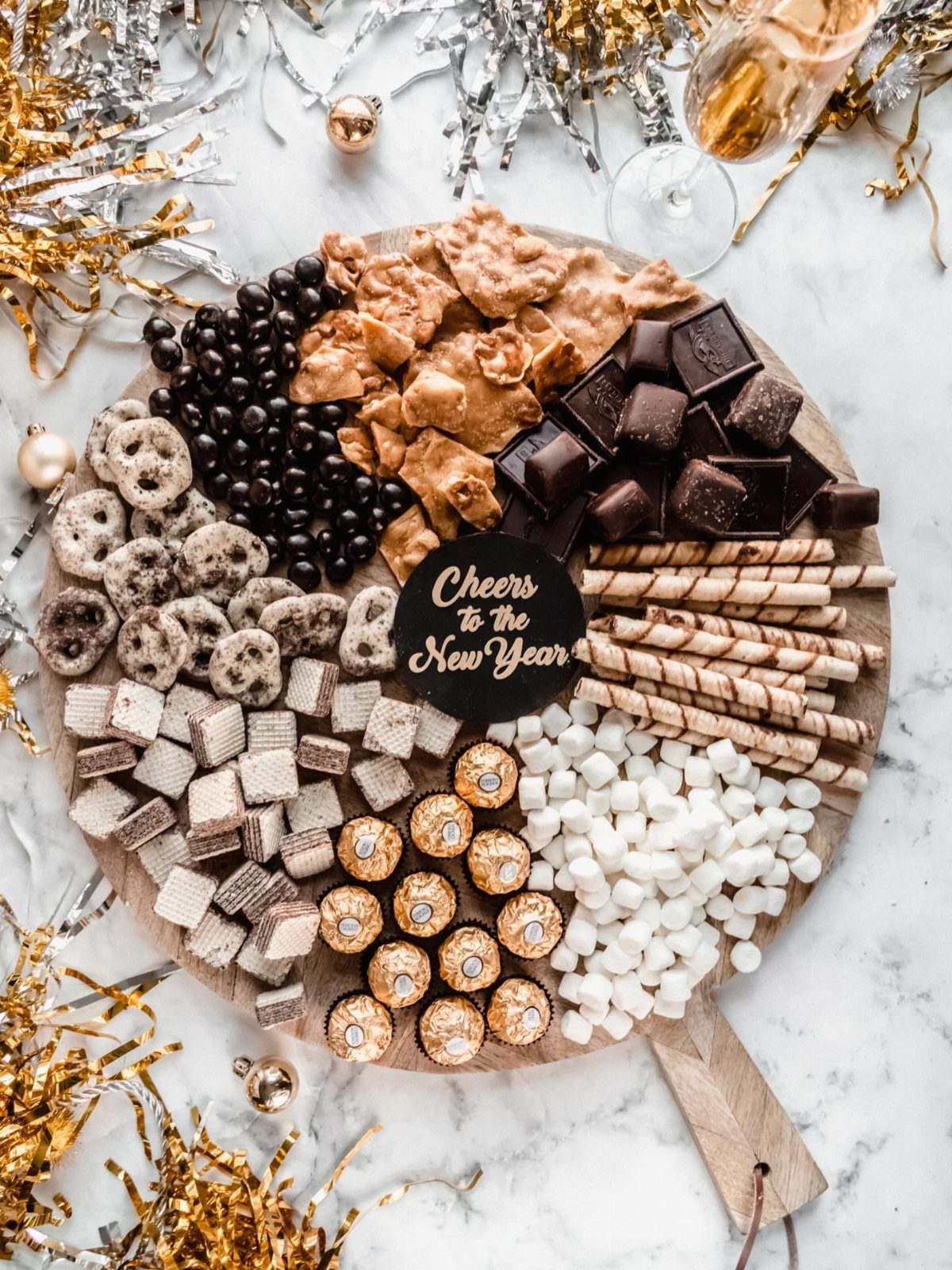 Dessert board with chocolates and marshmallows surrounded by gold and silver New Year tinsel.