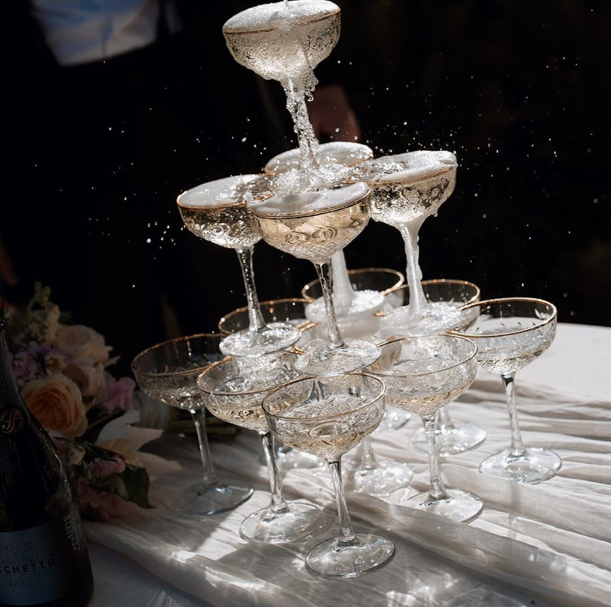 Champagne tower of gold-rimmed coupe glasses overflowing with sparkling wine on white linen.