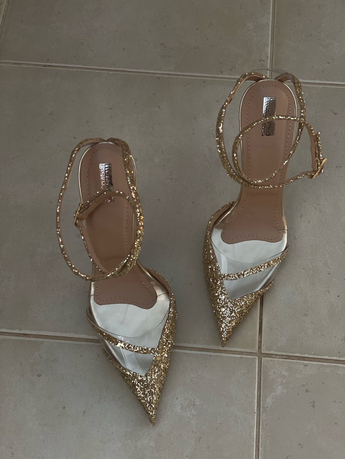 Gold glittery high-heeled sandals with ankle straps on a grey tiled floor.